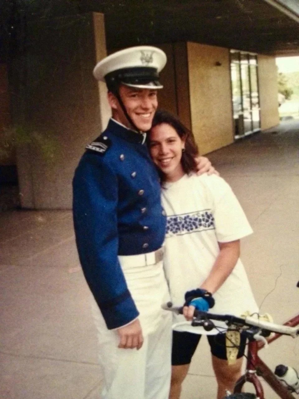 A man dressed as a U.S. Marine in uniform and a woman smiling, holding a bicycle, standing outdoors.